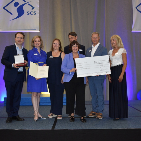 Sechs Personen auf einer B&uuml;hne halten Auszeichnungen und einen gro&szlig;en Scheck. Im Hintergrund Banner mit dem Logo &bdquo;SCS&ldquo;.