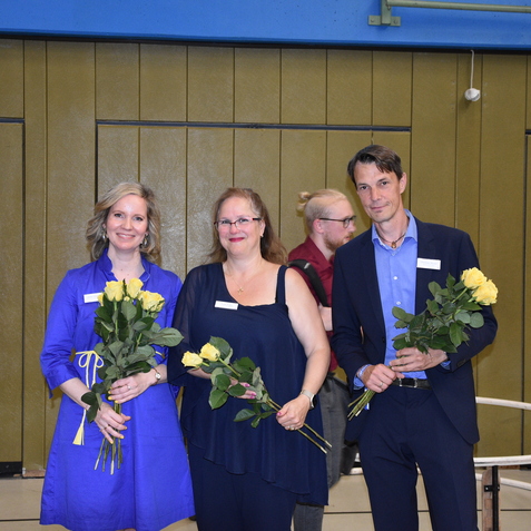 Drei Personen stehen in einer Sporthalle und halten jeweils einen Strau&szlig; gelber Rosen in der Hand.
