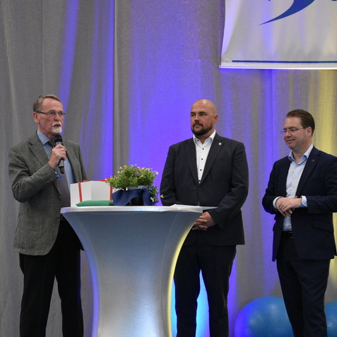 Ein rednerisches Ereignis mit drei Männern am Podium, einer spricht ins Mikrofon, im Hintergrund sind blaue Dekorationen.