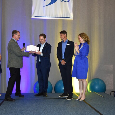 Zwei Männer überreichen sich ein Buch auf einer Bühne, umgeben von applaudierenden Personen in formalem Outfit.