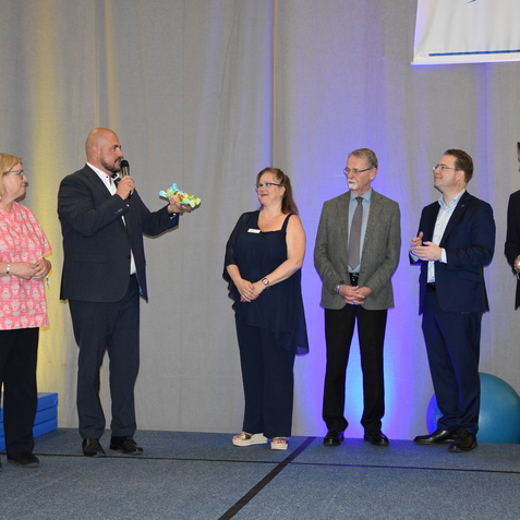 Gruppenvorstellung auf einer Bühne, während ein Mann mit Mikrofon eine Auszeichnung überreicht. Das Publikum schaut gespannt zu.