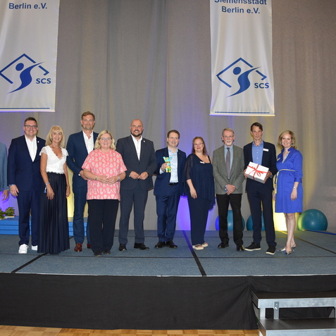 Gruppe von Menschen auf einer Bühne bei einer Veranstaltung, im Hintergrund Banner der SCS Berlin e.V. sichtbar.