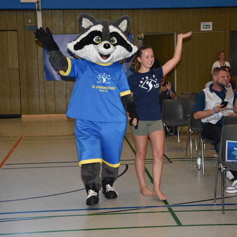 Maskottchen in blauem Outfit begleitet ein Mädchen auf einer Sportveranstaltung, Publikum applaudiert im Hintergrund.