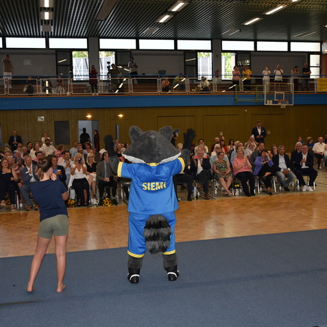 Maskottchen in blauer Sportkleidung mit „SIEM“ auf dem Rücken steht vor einem applaudierenden Publikum in einer Sporthalle.