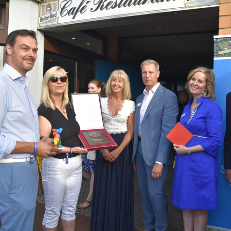 Gruppe von f&uuml;nf Personen vor einem Caf&eacute;, eine Frau h&auml;lt eine Auszeichnung und eine farbige Figur in der Hand.