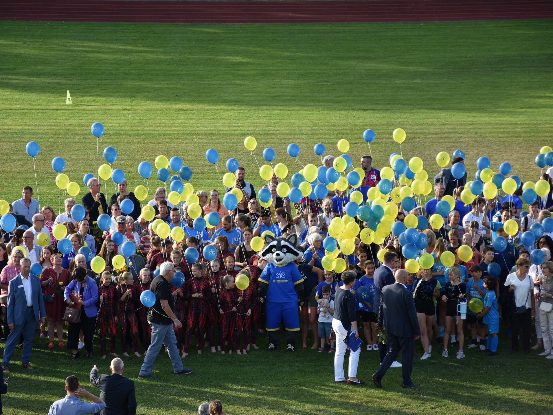 Gruppenszene auf einem Rasen mit Menschen, die gelbe und blaue Luftballons halten, während ein Maskottchen in der Mitte steht.
