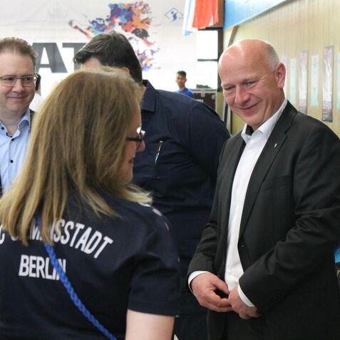 Zwei M&auml;nner in Anz&uuml;gen unterhalten sich mit einer Frau in Sportkleidung bei einer Veranstaltung in Berlin.