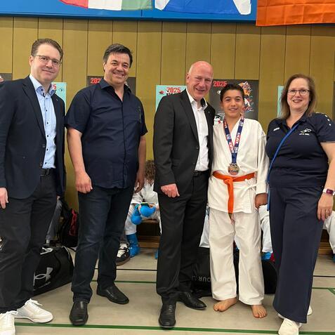 F&uuml;nf Personen stehen l&auml;chelnd zusammen, ein junger K&auml;mpfer in Karate-Duniform mit Medaille und orangefarbener G&uuml;rteld.