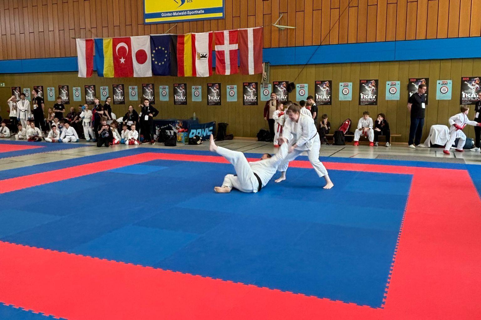 Zwei Kampfsportler in Wei&szlig;en Anz&uuml;gen trainieren auf einer blauen und roten Matte in einer Sporthalle mit Flaggen.