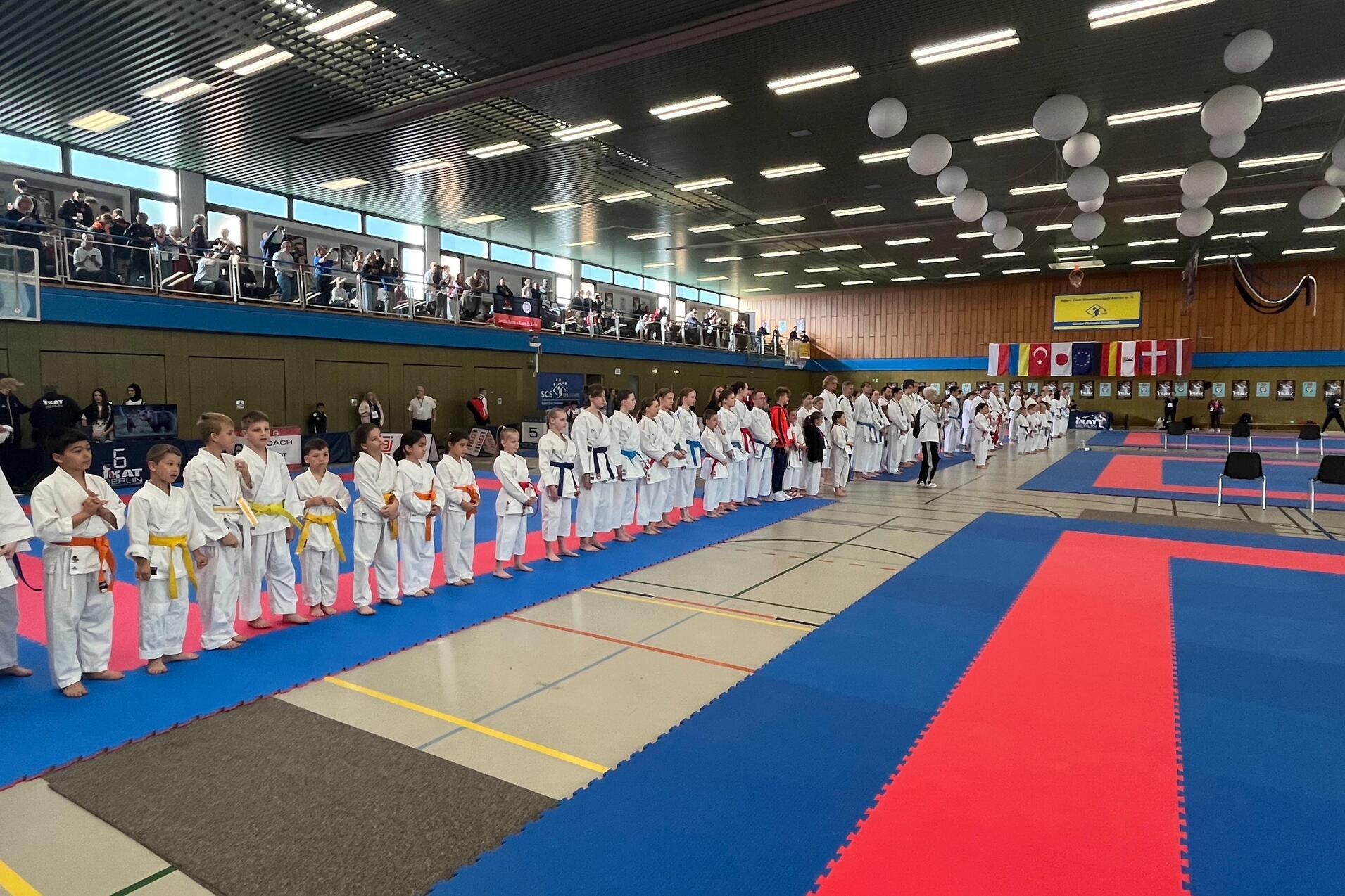 Karateka in wei&szlig;en Anz&uuml;gen stehen in Reihen auf blauer und roter Matte, Zuschauer sitzen an der Trib&uuml;ne im Hintergrund.