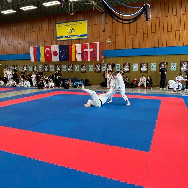Zwei Kampfsportler in Wei&szlig;en Anz&uuml;gen trainieren auf einer blauen und roten Matte in einer Sporthalle mit Flaggen.