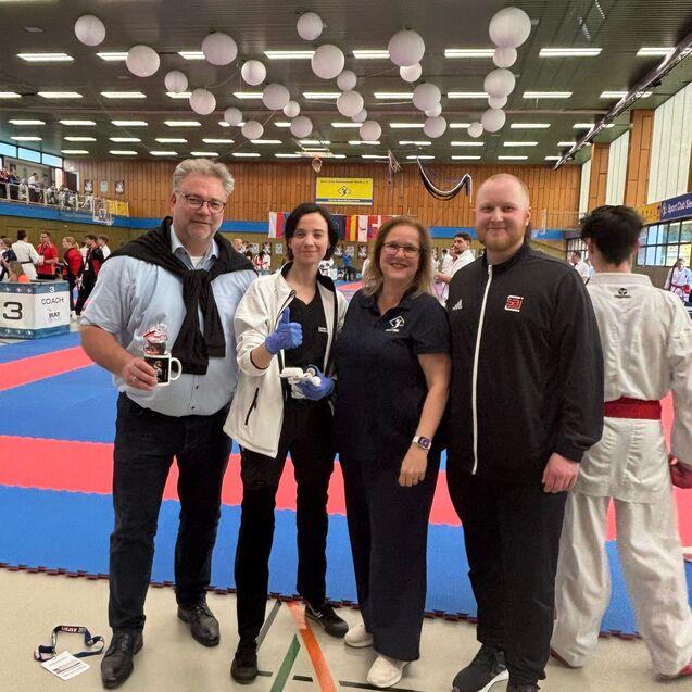 Vier Personen stehen in einem Sportzentrum neben einer Wettkampfbahn f&uuml;r Karate, Teilnehmer im Hintergrund.