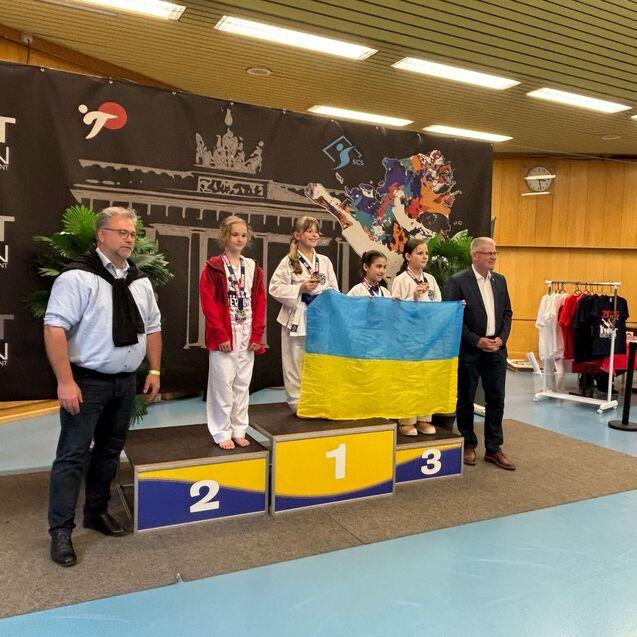 Drei Medaillengewinnerinnen im Karate stehen auf dem Podium, eine h&auml;lt die ukrainische Flagge, ein Mann steht daneben.