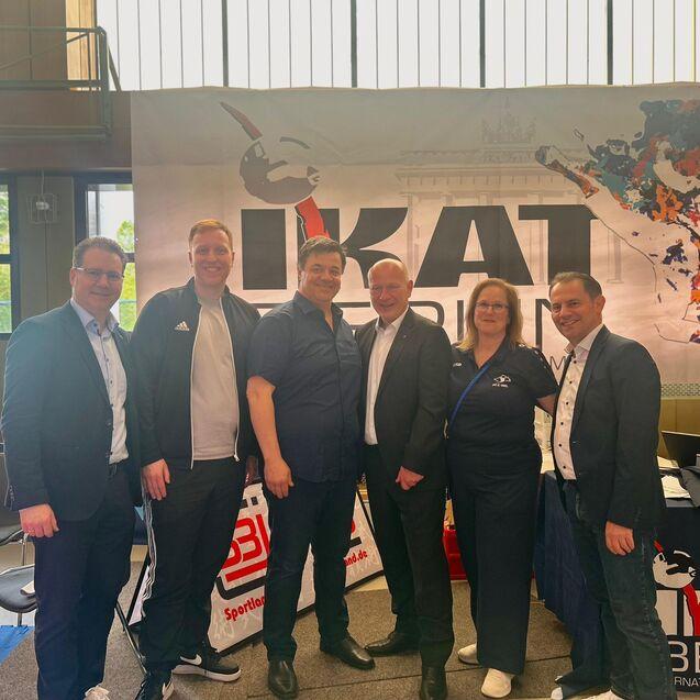 Sechs Personen in sportlicher Kleidung stehen vor einem Banner mit der Aufschrift &bdquo;IKAT Berlin&ldquo; in einer Sporthalle.