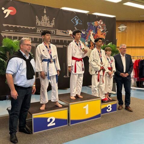 Vier Jugendliche im Karateanzug stehen auf dem Treppchen, Medaillen um den Hals, während zwei Erwachsene daneben posieren.