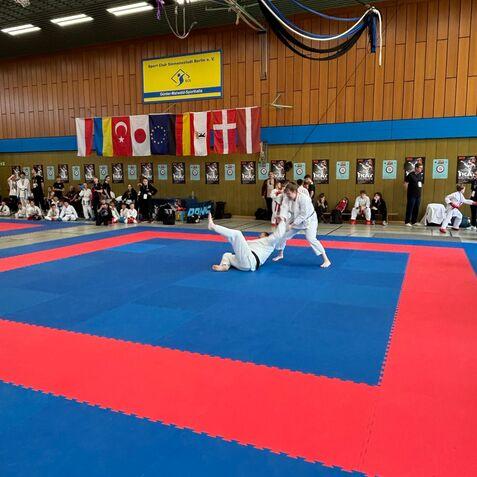 Zwei Kampfsportler in Weißen Anzügen trainieren auf einer blauen und roten Matte in einer Sporthalle mit Flaggen.
