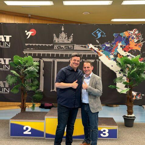 Zwei Männer stehen lächelnd vor einem großen Banner mit dem Brandenburger Tor und dem Logo des IKAT Berlin.