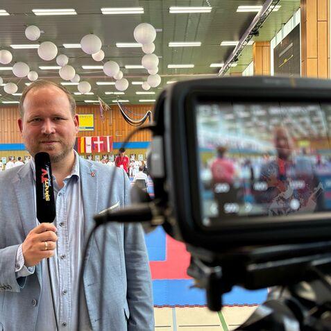 Mann mit Mikrofon steht vor einer Sportarena mit Kampfsportlern im Hintergrund, gefilmt durch eine Kamera.