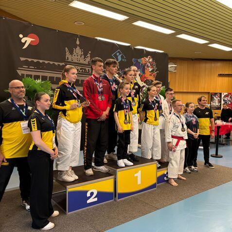 Sieben Sportler stehen auf dem Siegerpodest, gekleidet in Karateanzüge und Medaillen, während Zuschauer im Hintergrund stehen.