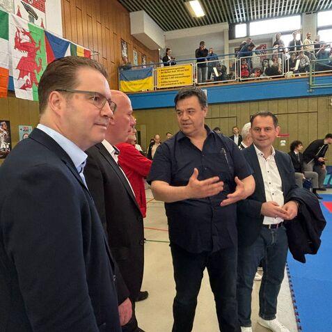 Vier Personen in Anzügen stehen in einer Sporthalle, während sie sich angeregt unterhalten. Im Hintergrund sind Zuschauer zu sehen.