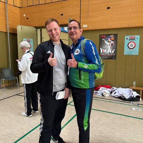 Zwei Männer geben Daumen hoch, während im Hintergrund eine Person mit Kamera steht. Plakate sind an der Wand sichtbar.