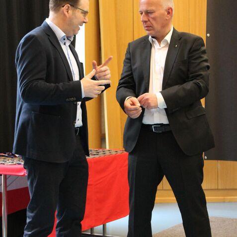 Zwei Männer im Gespräch, einer in Anzug und der andere in legerer Kleidung, vor einem Tisch mit rotem Tuch im Hintergrund.