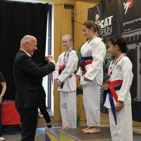 Drei Kinder in Karateanzügen stehen auf dem Podium, während ein Mann ihnen Medaillen überreicht.