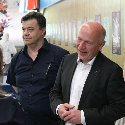 Zwei Männer im Gespräch, während ein Junge mit Medaille neugierig zuhört, in einer sportlichen Veranstaltungsumgebung.