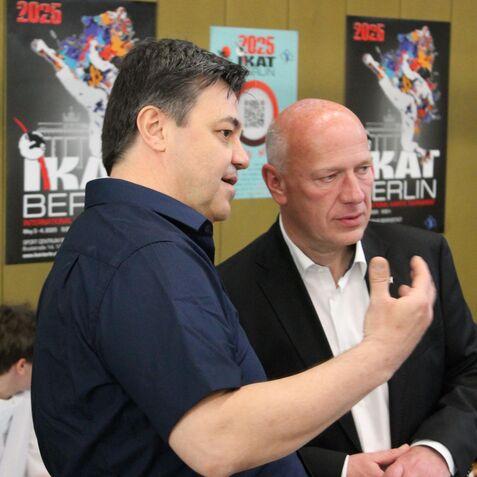 Zwei Männer im Gespräch vor bunten Plakaten, einer trägt ein schwarzes, der andere ein weißes Hemd.