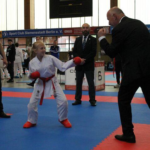 Mädchen im Karateanzug mit rotem Gürtel und Boxhandschuhen steht auf einer Wettkampfmatte und bereitet sich auf einen Angriff vor.