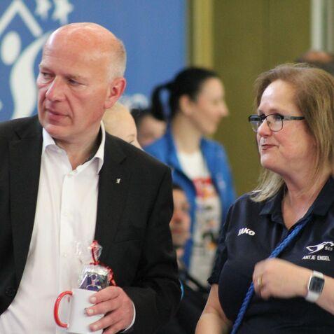 Zwei Personen unterhalten sich in einer Sporthalle, eine mit Brille und einer Tasse, der andere im Anzug.