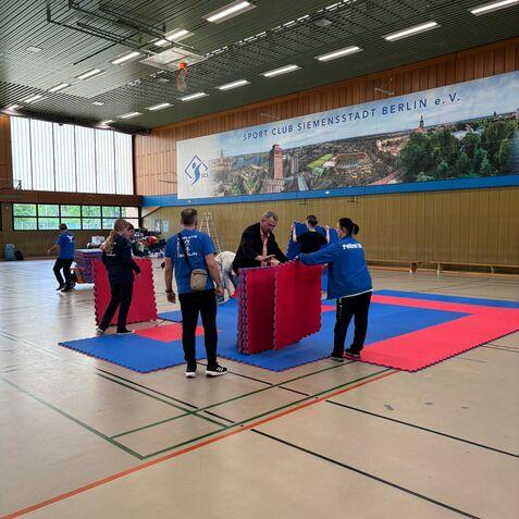 Gruppen von Menschen in blauen T-Shirts stellen Matten und Schutzeinrichtungen in einer Sporthalle auf.