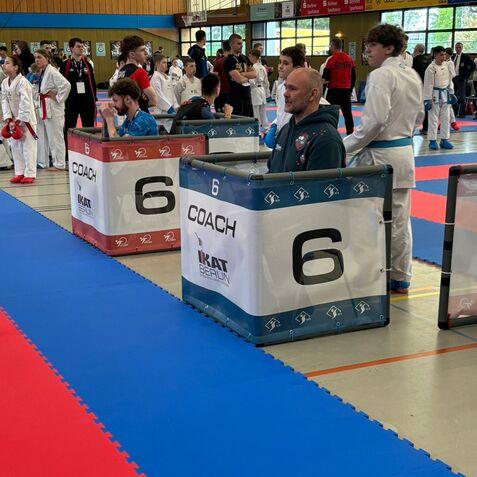 Mehrere Jugendliche und Trainer sitzen und stehen auf einer Matte in einem Trainingsraum während eines Karate-Wettkampfs.