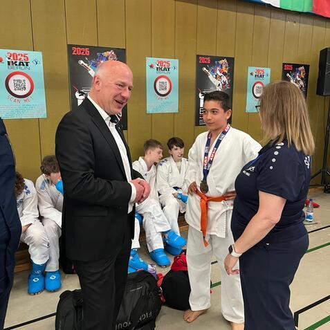 Gespräch zwischen einem Mann in Anzug und einem jungen Karateka mit Medaille, umgeben von weiteren Kindern in Judogis.