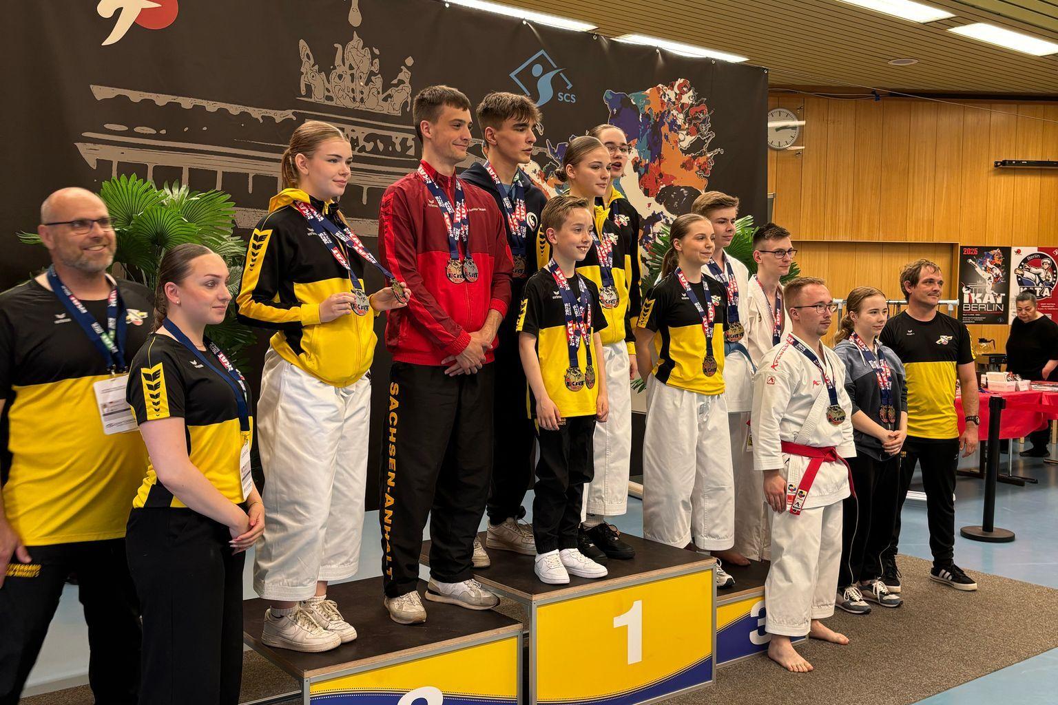 Sieben Sportler stehen auf dem Siegerpodest, gekleidet in Karateanz&uuml;ge und Medaillen, w&auml;hrend Zuschauer im Hintergrund stehen.