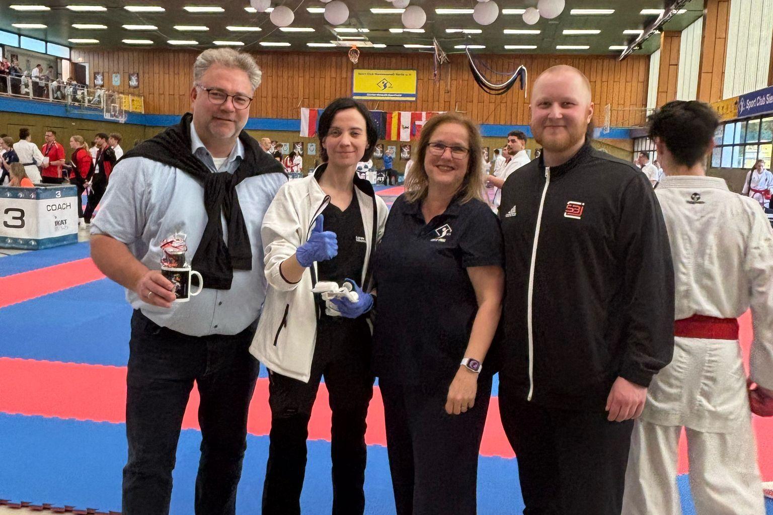 Vier Personen stehen in einem Sportzentrum neben einer Wettkampfbahn f&uuml;r Karate, Teilnehmer im Hintergrund.