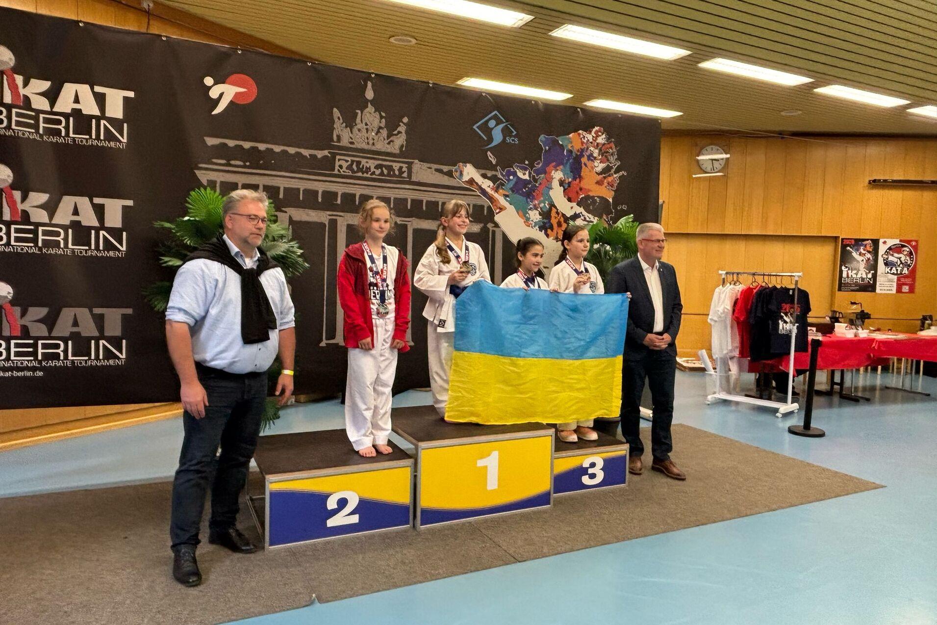 Drei Medaillengewinnerinnen im Karate stehen auf dem Podium, eine h&auml;lt die ukrainische Flagge, ein Mann steht daneben.