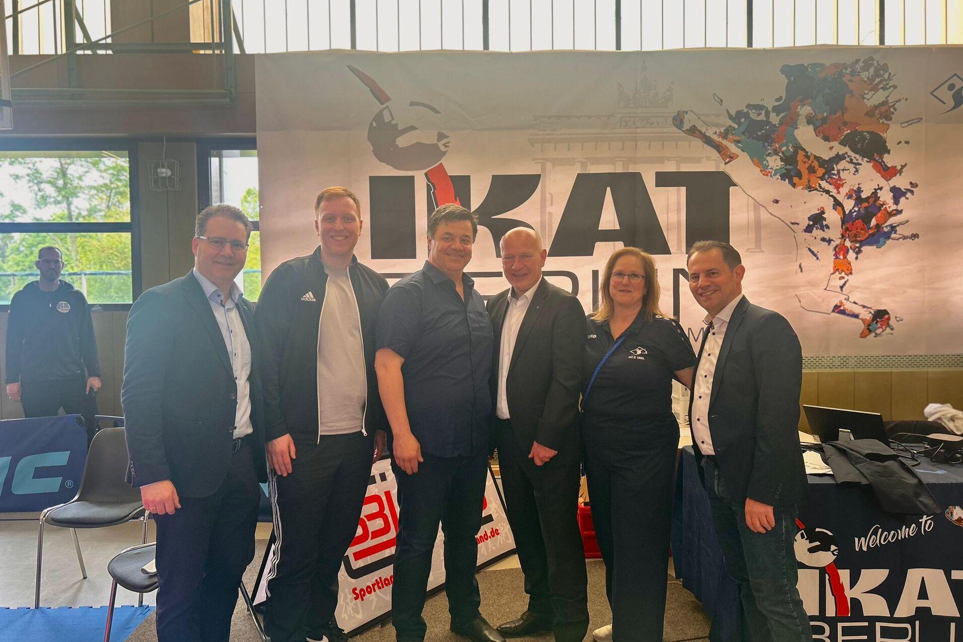 Sechs Personen in sportlicher Kleidung stehen vor einem Banner mit der Aufschrift &bdquo;IKAT Berlin&ldquo; in einer Sporthalle.