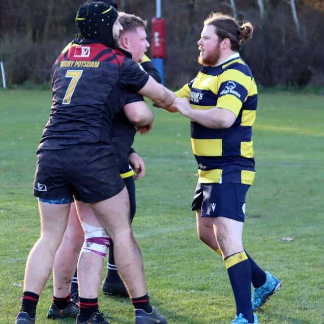 Zwei Rugbyspieler in Kontakt, w&auml;hrend ein dritter Spieler mit einem gepolsterten Helm beobachtet. Rasenplatz im Hintergrund.