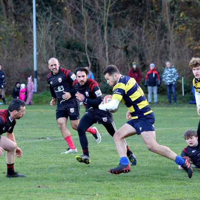 Rugby-Spieler in gelb-schwarz tr&auml;gt den Ball, w&auml;hrend andere Spieler ihn verfolgen, im Hintergrund Zuschauer auf einem Sportfeld.