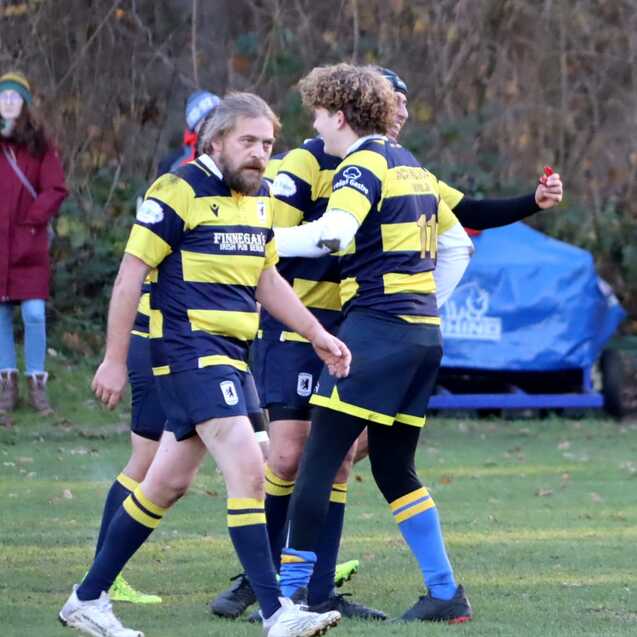 Zwei Rugbyspieler in gestreiften Trikots stehen auf einem Sportplatz, w&auml;hrend ein Zuschauer im Hintergrund steht.