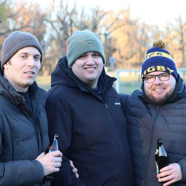 Drei M&auml;nner stehen l&auml;chelnd auf einem Sportplatz und halten Flaschen in den H&auml;nden, umgeben von B&auml;umen.