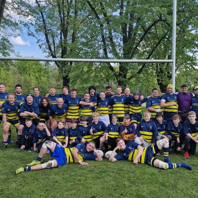 Gruppenfoto einer Rugbymannschaft bestehend aus Erwachsenen und Kindern auf einer Wiese, mit einem Rugbytor im Hintergrund.