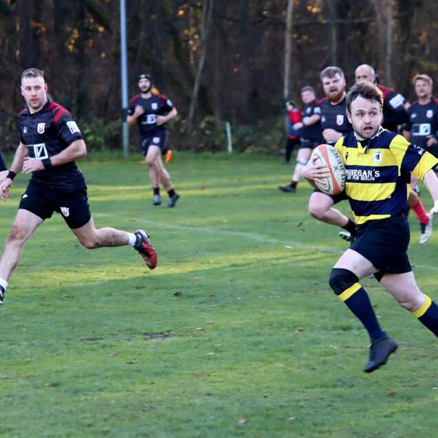 Rugbyspieler in gelb-schwarzer Uniform sprintet mit einem Ball &uuml;ber das Spielfeld, w&auml;hrend andere Spieler verfolgen.