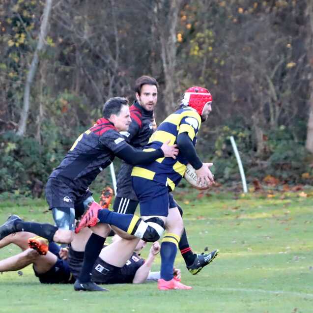 Drei Rugbyspieler in Aktion auf einem Spielfeld; ein Spieler tr&auml;gt einen roten Helm und h&auml;lt den Ball.