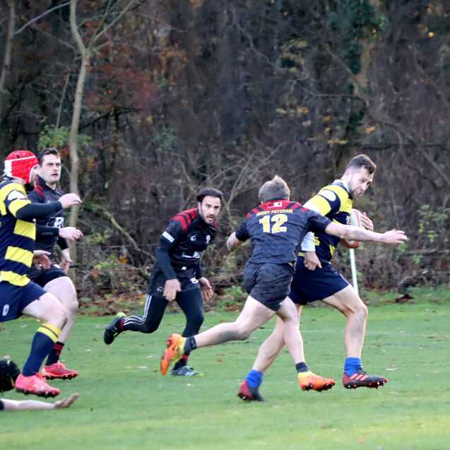 Zwei Rugbyspieler laufen auf einem verdeckten Spielfeld, w&auml;hrend andere Spieler im Hintergrund sich positionieren.