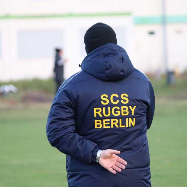 Person mit einer blauen Jacke mit der Aufschrift "SCS RUGBY BERLIN" steht auf einem Rugbyfeld und beobachtet das Geschehen.