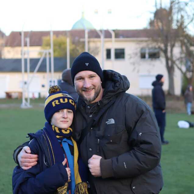 Ein Junge mit Rugby-Schal und eine Person in winterlicher Kleidung stehen l&auml;chelnd auf einem Sportplatz.