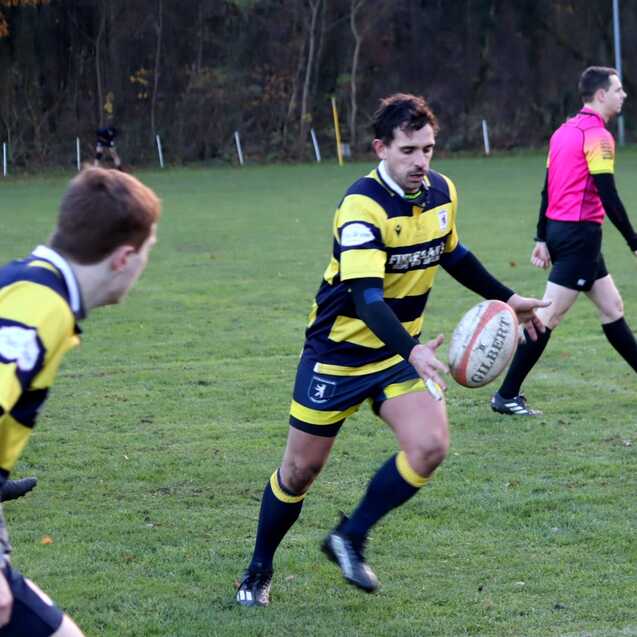 Rugby-Spieler in gestreifter Kleidung f&auml;ngt einen Ball auf einem gr&uuml;nen Spielfeld, w&auml;hrend ein Schiedsrichter zusieht.