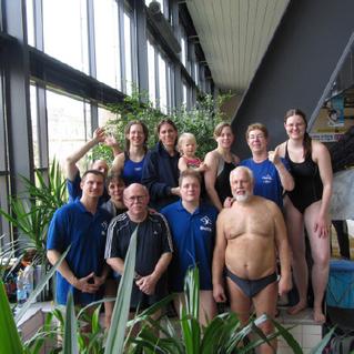 Gruppennahaufnahme von Schwimmern in Badebekleidung, stehend und lachend neben einem Innenpool. Pflanzen im Hintergrund.